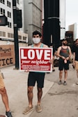 A person wearing a face mask holds up a sign that reads 'LOVE your neighbor' followed by a list of diverse groups such as black, brown, immigrant, disabled, religiously different, LGBTQ. They are standing on a city sidewalk surrounded by tall buildings. Other people are present in the background, some possibly holding other signs.