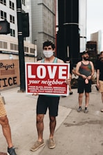 A person wearing a face mask holds up a sign that reads 'LOVE your neighbor' followed by a list of diverse groups such as black, brown, immigrant, disabled, religiously different, LGBTQ. They are standing on a city sidewalk surrounded by tall buildings. Other people are present in the background, some possibly holding other signs.