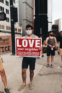 A person wearing a face mask holds up a sign that reads 'LOVE your neighbor' followed by a list of diverse groups such as black, brown, immigrant, disabled, religiously different, LGBTQ. They are standing on a city sidewalk surrounded by tall buildings. Other people are present in the background, some possibly holding other signs.