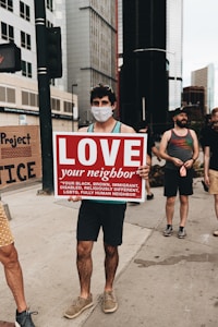 A person wearing a face mask holds up a sign that reads 'LOVE your neighbor' followed by a list of diverse groups such as black, brown, immigrant, disabled, religiously different, LGBTQ. They are standing on a city sidewalk surrounded by tall buildings. Other people are present in the background, some possibly holding other signs.