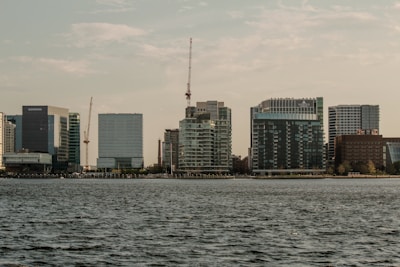 A cityscape featuring a waterfront view with several modern high-rise buildings. The skyline includes construction cranes and high-rise apartments with a body of water in the foreground, under a partly cloudy sky.
