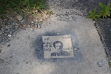 A stencil-style artwork is painted on a concrete surface, featuring a person's face and the text 'I'm kind of a big deal'. Surrounding the artwork, there are patches of grass, small rocks, and more concrete.