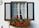 A charming window with partially open green shutters is adorned with delicate white curtains. A vibrant window box filled with lush green leaves and bright red flowers adds a splash of color to the scene. The white wall background complements the simplicity and elegance of the window design.