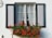 A charming window with partially open green shutters is adorned with delicate white curtains. A vibrant window box filled with lush green leaves and bright red flowers adds a splash of color to the scene. The white wall background complements the simplicity and elegance of the window design.