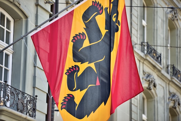 A vibrant flag featuring a bold black bear with red claws and tongue on a split red and yellow background is hanging in front of an ornate building facade. The structure has decorative elements, wrought iron railings, and large windows.