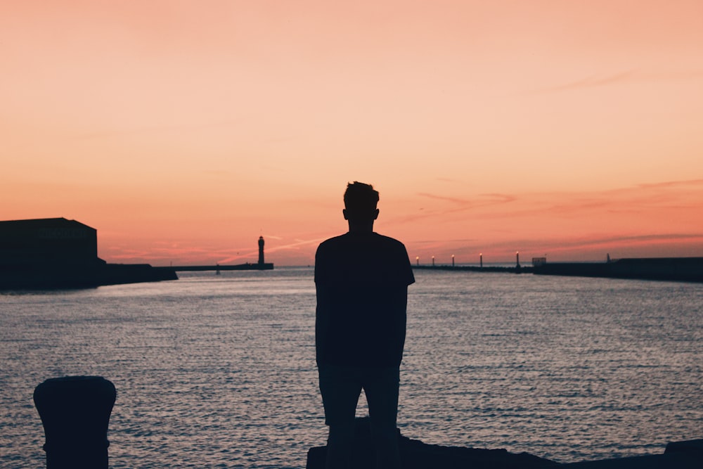 silhouette of man standing on sea shore during sunset