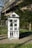 A utility box painted to resemble a house with windows and doors, featuring silhouettes of people in the lower sections. Surrounding it is a grassy area with daisies, and behind is a modern building partially hidden by trees.