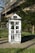 A utility box painted to resemble a house with windows and doors, featuring silhouettes of people in the lower sections. Surrounding it is a grassy area with daisies, and behind is a modern building partially hidden by trees.