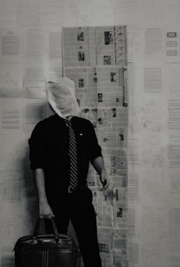 A person stands in a room with a wall covered in newspapers and documents. They are wearing a black shirt, a tie, and holding a briefcase. The individual's head is obscured by a cloth bag.