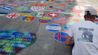 A man wearing a cap and a T-shirt with a printed image is kneeling on a pavement decorated with colorful circles, each representing a different country's flag. In the foreground, a heart shape is painted with a message that promotes unity amongst different religions, languages, and skin colors.
