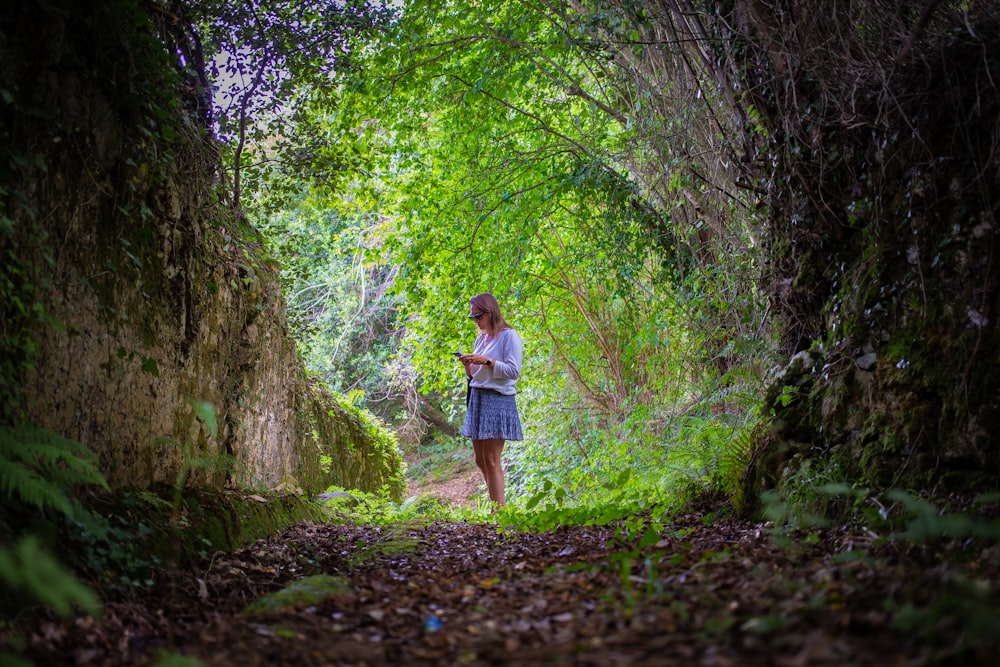 woman in white shirt and blue skirt walking on pathway between green trees during daytime