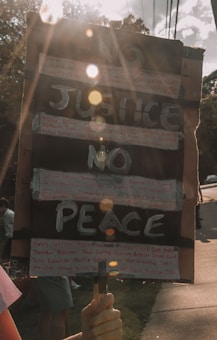 A hand-held protest sign with the words 'NO JUSTICE NO PEACE' written in large, bold letters. Additional smaller text, possibly names, is handwritten around the main message. The sign is held by a person, and sunlight creates lens flares across the image, adding a dramatic effect. The background shows trees and a sidewalk with a few people present, partially out of focus.