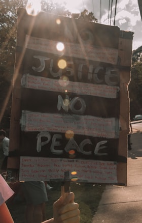 A hand-held protest sign with the words 'NO JUSTICE NO PEACE' written in large, bold letters. Additional smaller text, possibly names, is handwritten around the main message. The sign is held by a person, and sunlight creates lens flares across the image, adding a dramatic effect. The background shows trees and a sidewalk with a few people present, partially out of focus.