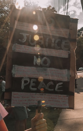 A hand-held protest sign with the words 'NO JUSTICE NO PEACE' written in large, bold letters. Additional smaller text, possibly names, is handwritten around the main message. The sign is held by a person, and sunlight creates lens flares across the image, adding a dramatic effect. The background shows trees and a sidewalk with a few people present, partially out of focus.