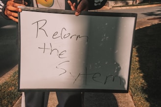 A person holding a white board with handwritten text saying 'Reform the System'. The board is partially overshadowed and is being held with both hands.
