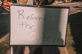 A person holding a white board with handwritten text saying 'Reform the System'. The board is partially overshadowed and is being held with both hands.