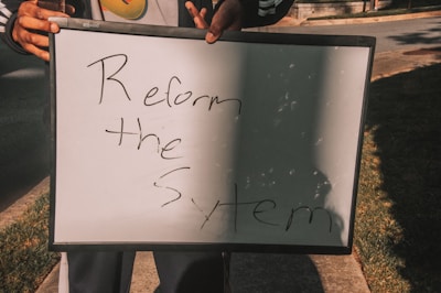 A person holding a white board with handwritten text saying 'Reform the System'. The board is partially overshadowed and is being held with both hands.