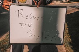 A person holding a white board with handwritten text saying 'Reform the System'. The board is partially overshadowed and is being held with both hands.