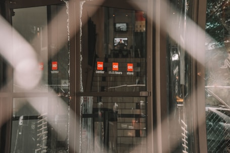 A glass entrance with logos displayed on the doors, reflecting an indoor space with stairs that appear in the background. The image has a soft blur effect, creating a sense of depth with a focus on the signage.