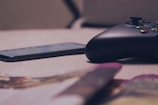 A close-up view of a gaming controller and a smartphone resting on a soft, beige surface. The setup is casually placed, with blurred objects in the foreground suggesting a relaxed, at-home environment.