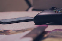 A close-up view of a gaming controller and a smartphone resting on a soft, beige surface. The setup is casually placed, with blurred objects in the foreground suggesting a relaxed, at-home environment.