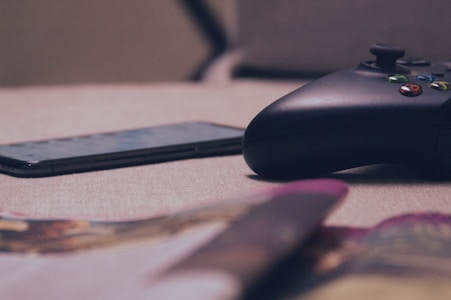 A close-up view of a gaming controller and a smartphone resting on a soft, beige surface. The setup is casually placed, with blurred objects in the foreground suggesting a relaxed, at-home environment.