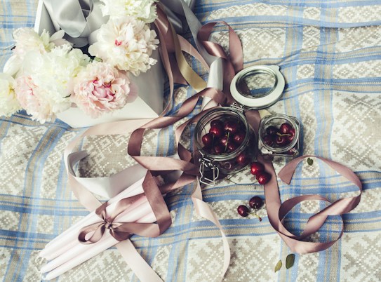 A still life arrangement featuring a collection of pink and white peonies placed in a decorative box. A set of pink and brown ribbons is artistically draped across a textured, patterned fabric surface with a blue checkered design. Glass jars filled with cherries, some scattered around, add a touch of color and contrast to the subtle pastel tones of the flowers and fabric.