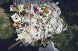 A picnic setup on a patterned blanket spread over grass, featuring a variety of items including a basket of cherries, apples, flowers, and a tray with pastries and cups of tea. There are books, magazines, and decorative items like candles and a small flower pot. Sunglasses and a striped skirt of a person are partially visible at the edge.