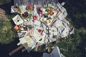 A picnic setup on a patterned blanket spread over grass, featuring a variety of items including a basket of cherries, apples, flowers, and a tray with pastries and cups of tea. There are books, magazines, and decorative items like candles and a small flower pot. Sunglasses and a striped skirt of a person are partially visible at the edge.
