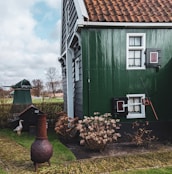 A charming rural scene featuring a traditional house with green wooden siding and white-framed windows. A rustic chimney pot is visible in the garden, surrounded by neatly trimmed shrubs and a cobblestone path. In the background, a small structure and a white bird are discernible, adding to the pastoral ambiance.