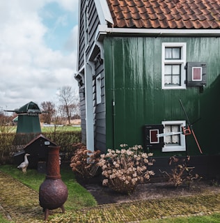 A charming rural scene featuring a traditional house with green wooden siding and white-framed windows. A rustic chimney pot is visible in the garden, surrounded by neatly trimmed shrubs and a cobblestone path. In the background, a small structure and a white bird are discernible, adding to the pastoral ambiance.