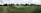 A panoramic view of a golf course featuring well-maintained grass, several sand bunkers, and a small pond. The foreground includes a paved path bordered by rope, leading through the green landscape. A modern clubhouse is situated on the right, surrounded by trees and open space.