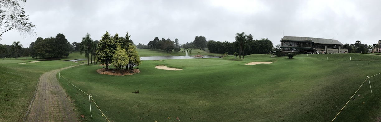 A panoramic view of a golf course featuring well-maintained grass, several sand bunkers, and a small pond. The foreground includes a paved path bordered by rope, leading through the green landscape. A modern clubhouse is situated on the right, surrounded by trees and open space.