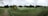 A panoramic view of a golf course featuring well-maintained grass, several sand bunkers, and a small pond. The foreground includes a paved path bordered by rope, leading through the green landscape. A modern clubhouse is situated on the right, surrounded by trees and open space.