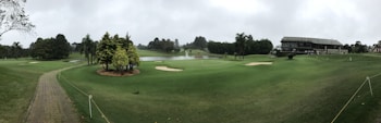A panoramic view of a golf course featuring well-maintained grass, several sand bunkers, and a small pond. The foreground includes a paved path bordered by rope, leading through the green landscape. A modern clubhouse is situated on the right, surrounded by trees and open space.