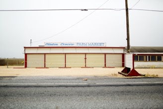 A closed roadside farm market with a faded sign sits on a gravel lot. The building is beige with red accents, featuring large garage-style doors. There is an empty road in the foreground and a utility pole with wires overhead.
