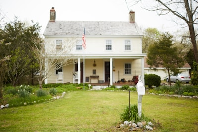 A two-story house with a white facade and a front porch, featuring an American flag on a flagpole. The house is surrounded by a well-maintained garden with grass and various shrubs and trees. There is a white mailbox in the foreground near the driveway, and a car is parked in the background.