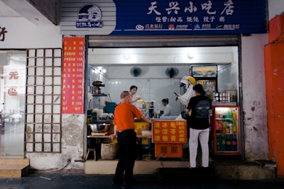A small street food shop with people ordering and preparing food. The storefront displays Chinese writing and price lists. Inside, a worker is preparing food, while a couple of customers wait at the counter. The shop has refrigerators displaying drinks and various utensils and appliances visible.