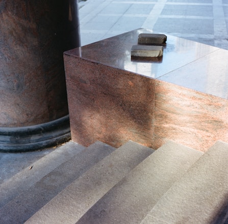 A close-up view of stone steps leading upward, adjacent to a polished, reddish-brown granite block with two stone bricks placed on top. A large cylindrical stone column sits nearby, with sunlight casting shadows on the smooth surfaces.
