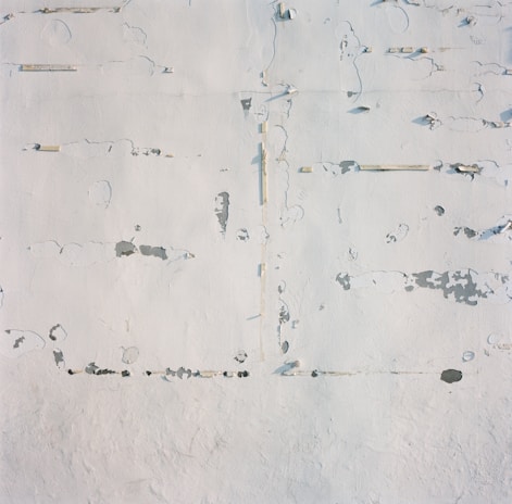 A textured surface that appears weathered, with patches of peeling paint and small cracks. The paint has faded in areas, revealing underlying layers. The surface is predominantly light in color, with natural imperfections creating an abstract pattern.