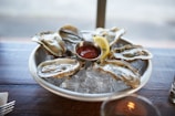 A platter of fresh oysters is served on a bed of ice, accompanied by a lemon wedge and a small dish of red sauce. The wooden table adds a rustic feel to the setting.