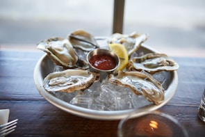 A platter of fresh oysters is served on a bed of ice, accompanied by a lemon wedge and a small dish of red sauce. The wooden table adds a rustic feel to the setting.