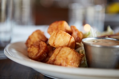 Golden-brown fried seafood pieces are arranged on a white plate, accompanied by a small metal cup filled with dipping sauce. The background is softly blurred, giving focus to the crispy texture of the seafood.
