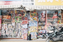 A storefront packed with an assortment of colorful toys, including action figures, toy cars, and various playsets, is displayed prominently. The shelves are stacked high with boxed toys and accessories, creating a vibrant and busy scene. A scooter is parked in front of the shop, adding to the bustling urban setting.