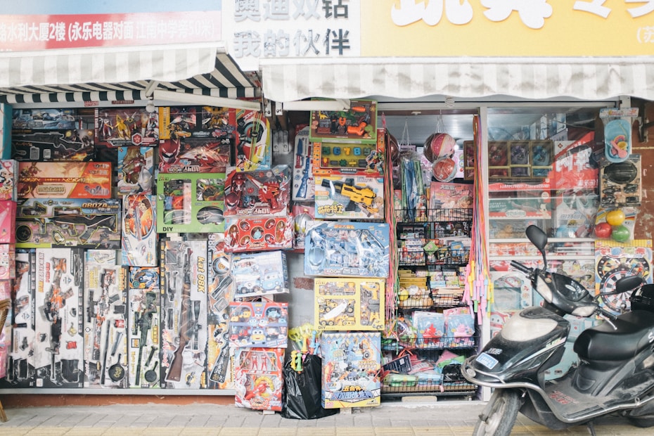 A storefront packed with an assortment of colorful toys, including action figures, toy cars, and various playsets, is displayed prominently. The shelves are stacked high with boxed toys and accessories, creating a vibrant and busy scene. A scooter is parked in front of the shop, adding to the bustling urban setting.
