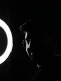 A silhouette of a person with glasses is partially illuminated by a circular light source on the left side.