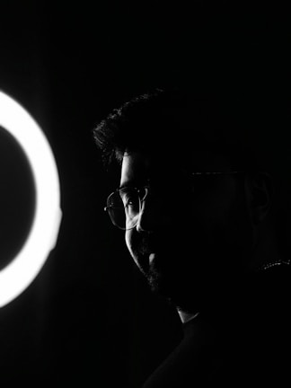 A silhouette of a person with glasses is partially illuminated by a circular light source on the left side.