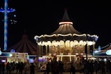 A brightly lit carousel is captured at night, surrounded by people and featuring classic ornate designs. The scene includes a large tent, possibly a circus or fairground tent, with multicolored lights illuminating the area. There are silhouettes of people gathered around, creating a lively atmosphere.