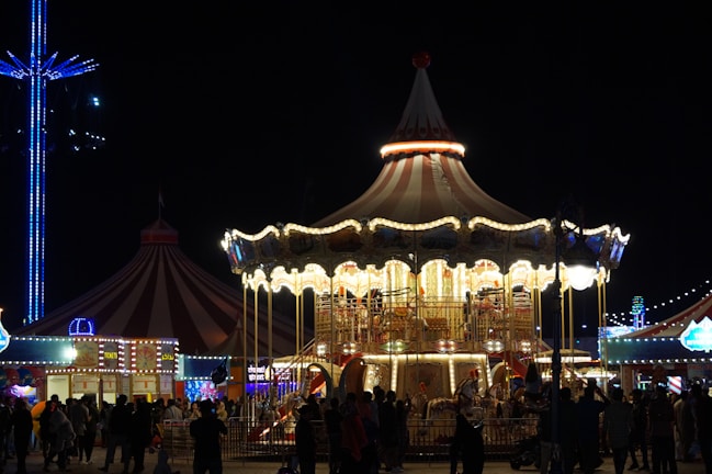 A brightly lit carousel is captured at night, surrounded by people and featuring classic ornate designs. The scene includes a large tent, possibly a circus or fairground tent, with multicolored lights illuminating the area. There are silhouettes of people gathered around, creating a lively atmosphere.