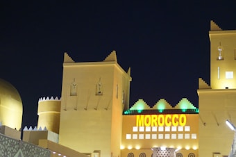 A Moroccan-themed building is illuminated at night with bright yellow lighting on traditional architecture featuring crenelated walls and towers. A prominent sign displaying the word 'MOROCCO' in bold yellow letters is visible.